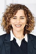 Frau mit lockigem, braunem Haar lächelt in die Kamera. Sie trägt einen Blazer über einem Pullover vor hellem Hintergrund.
