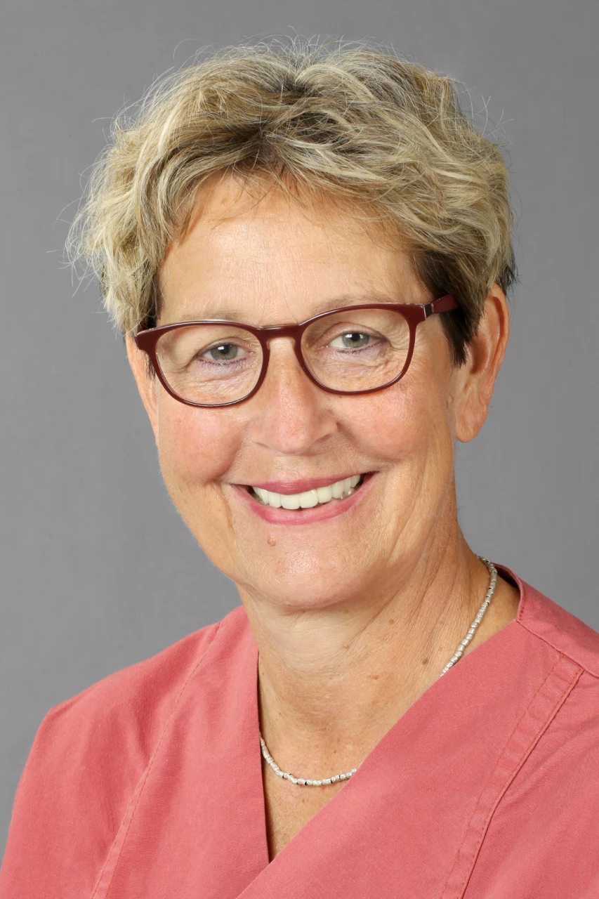 Porträt einer lächelnden Frau mit Kurzhaarschnitt, Brille und rosa Bluse vor grauem Hintergrund.