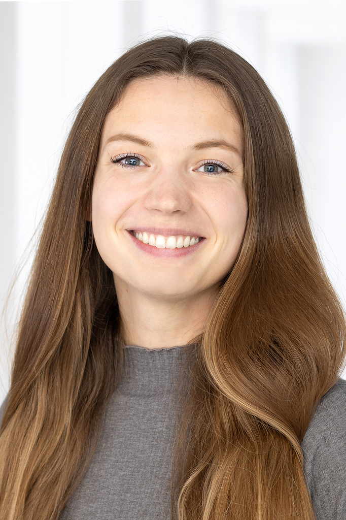 Lächelnde Frau mit langem braunen Haar, trägt einen grauen Pullover. Weißer Hintergrund.