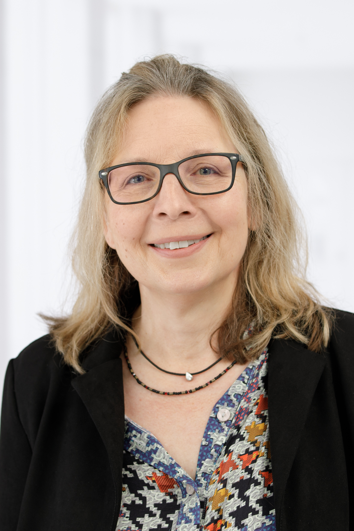 Frau mit Brille und langen Haaren lächelt vor neutralem Hintergrund, trägt bunte Bluse und schwarzen Blazer.