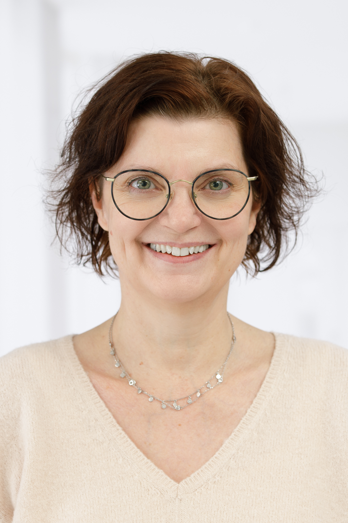 Frau mit braunem, lockigem Haar und Brille lächelt in die Kamera vor einem hellen Hintergrund.